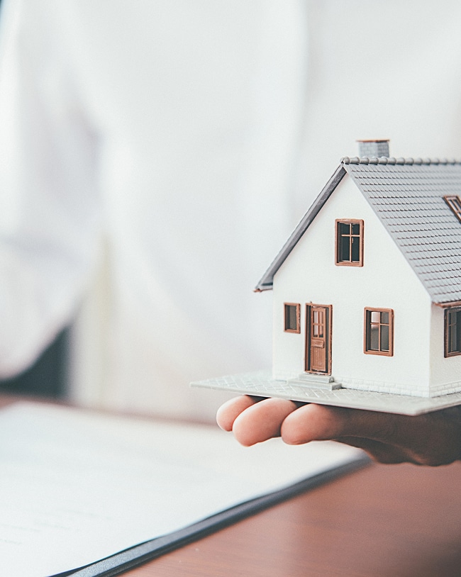 Achat immobilier: signer le contrat pour la maison de vos rêves. Main tenant une maquette de maison blanche avec un toit gris, posée au-dessus d'un document de contrat immobilier.